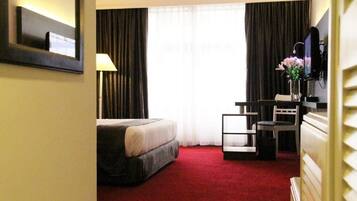 Une chambre d’hôtel avec un lit, un bureau, une chaise, une lampe, une télévision et une fenêtre avec des rideaux.