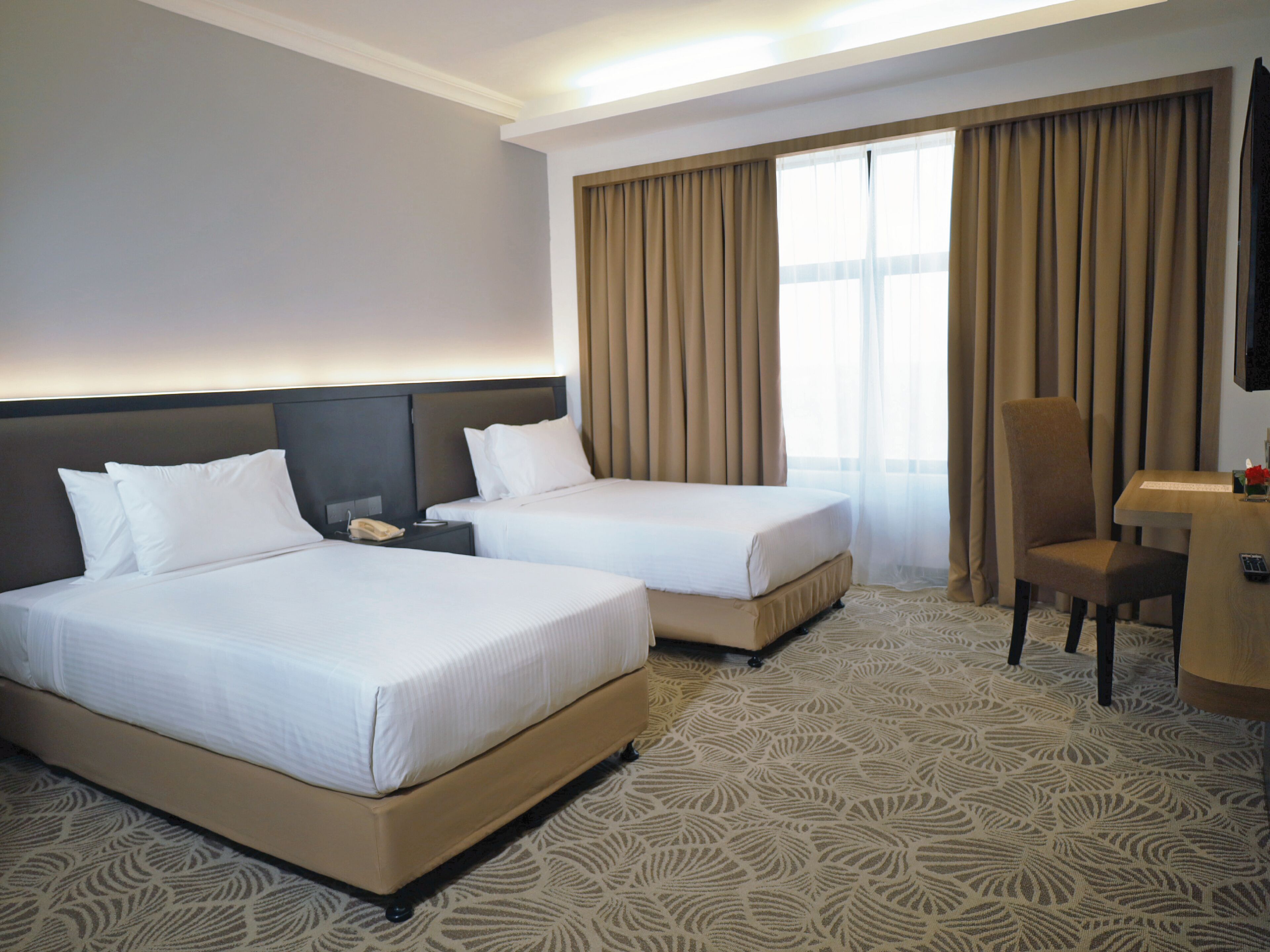Quarto de hotel com duas camas, uma escrivaninha, uma cadeira, telefone e uma janela grande com cortinas.
