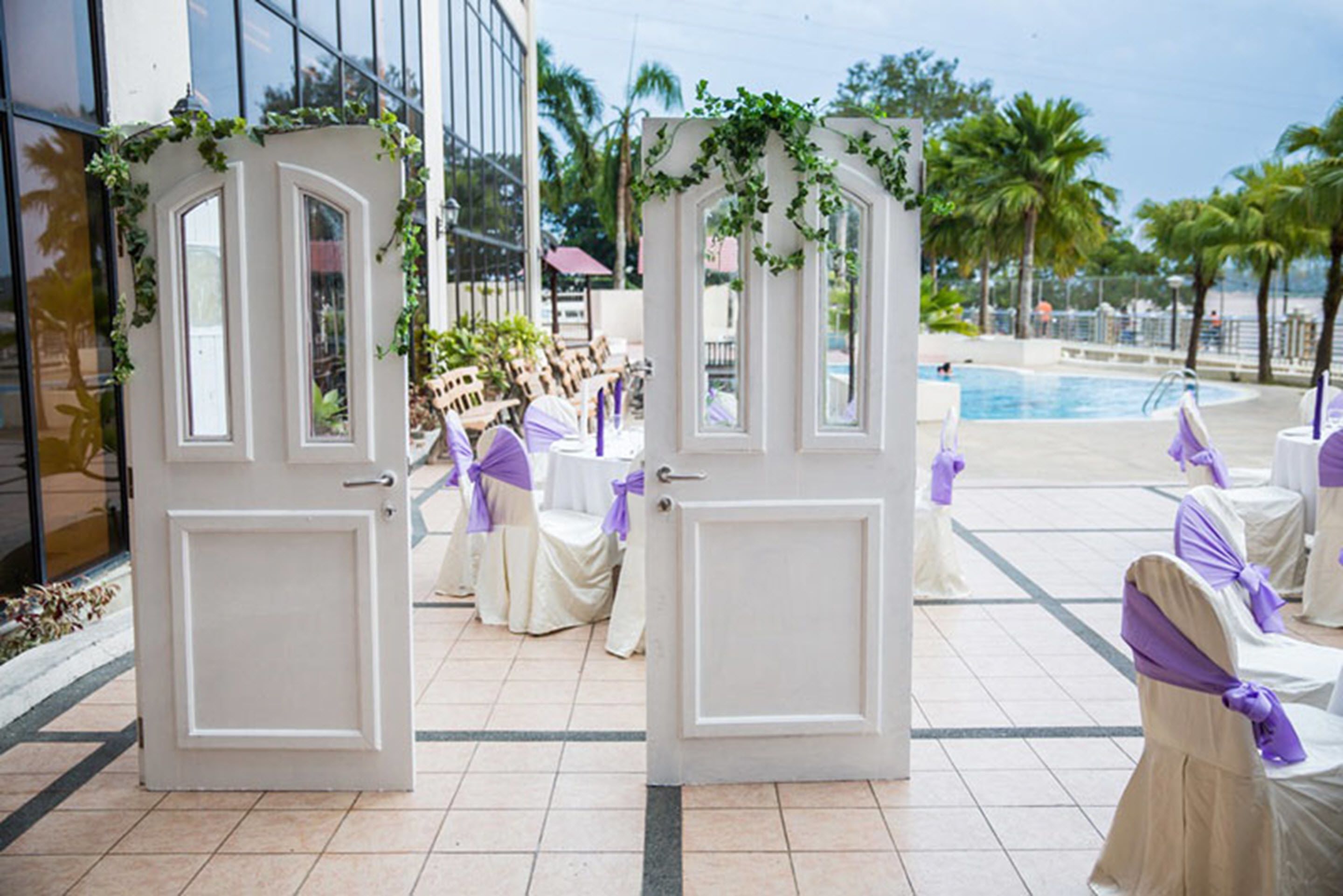 A hotel entrance with white doors, a pool area, and outdoor seating with purple sashes.