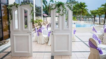 A hotel entrance with white doors, a pool area, and outdoor seating with purple sashes.