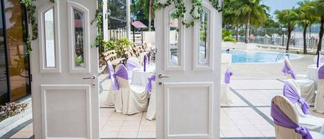 A hotel entrance with white doors, a pool area, and outdoor seating with purple sashes.