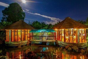 A resort with two thatched-roof structures connected by a glass bridge over a reflective water body, surrounded by lush greenery. - Poa Place Resort & Hotel - Where Nature Meets Comfort Eldoret (Eldoret)