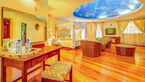 Une chambre d’hôtel spacieuse avec un grand lit, un coin salon avec des meubles en osier, une table à manger sur laquelle se trouve un plateau de bouteilles d’eau, et un plafond décoré de motifs nuageux.