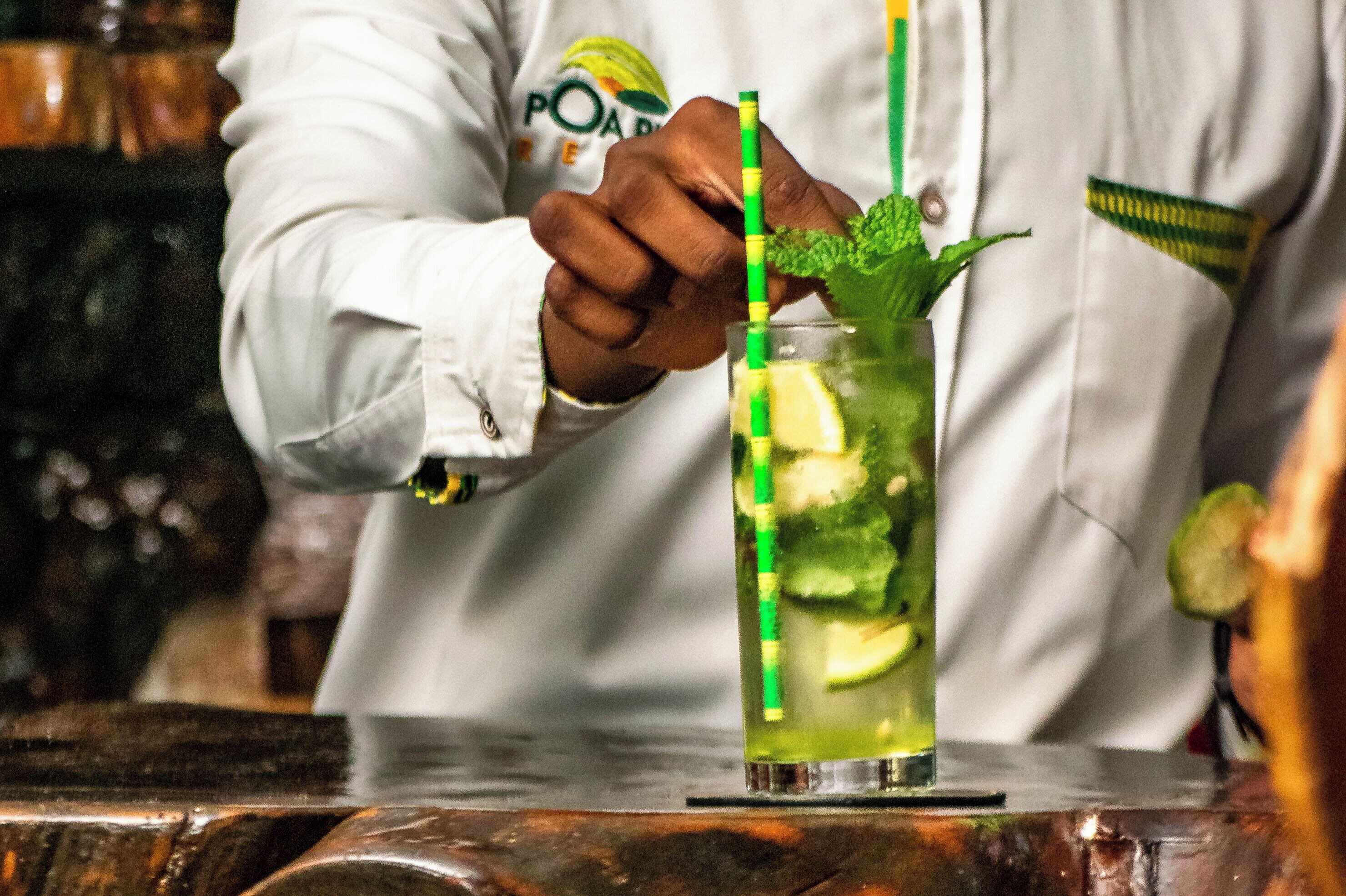 Une personne portant une chemise blanche prépare une boisson qu’elle agrémente d’une tranche de citron vert et de quelques feuilles de menthe.