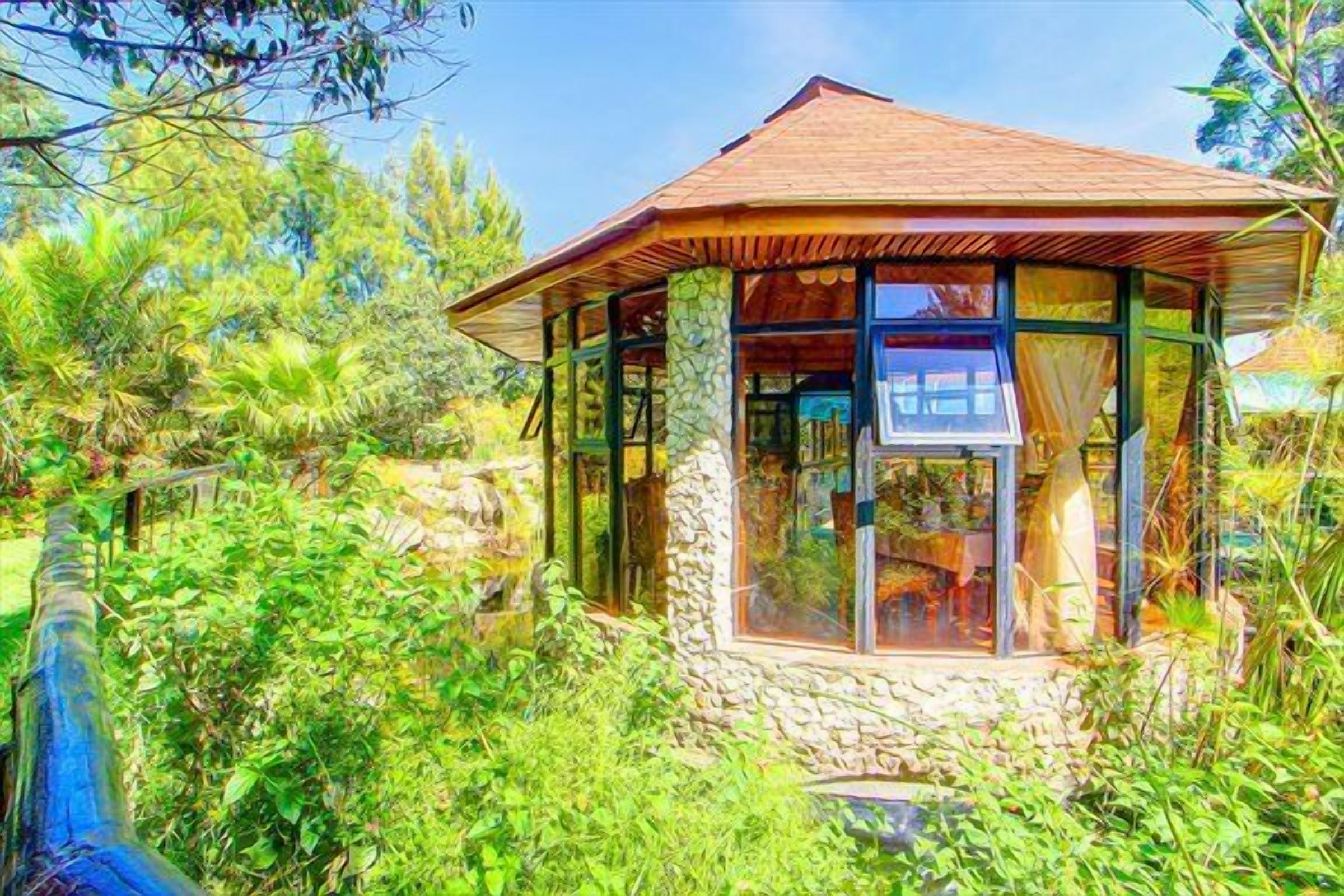 A small, stone-walled structure with a wooden roof and large windows, surrounded by lush greenery.