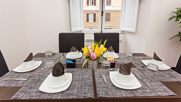 Mesa para cuatro lista para comer, con un florero, un mantel decorado y vistas de edificios a través de una ventana.