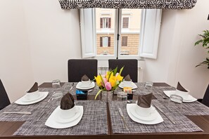 Mesa de jantar posta para quatro pessoas, com um vaso de flores, um jogo americano estampado e vista para edifícios através de uma janela.