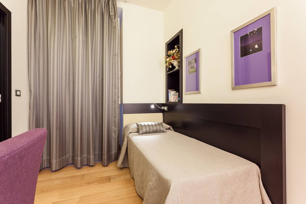 Un dormitorio pequeño y ordenado con una cama individual, una mesita de noche, una silla y una estantería de pared con adornos.