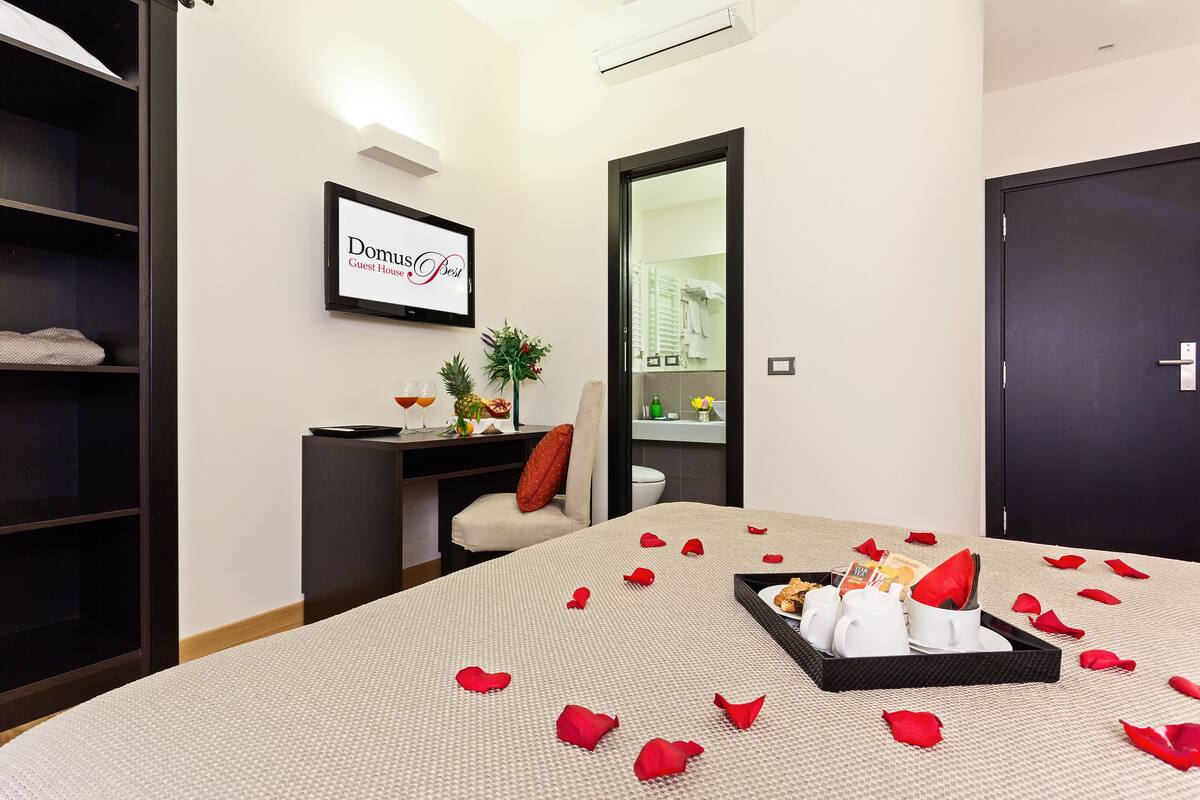 Habitación de hotel con cama, escritorio, silla y armario. Hay pétalos de rosa sobre la cama y una bandeja con toallas y un libro.