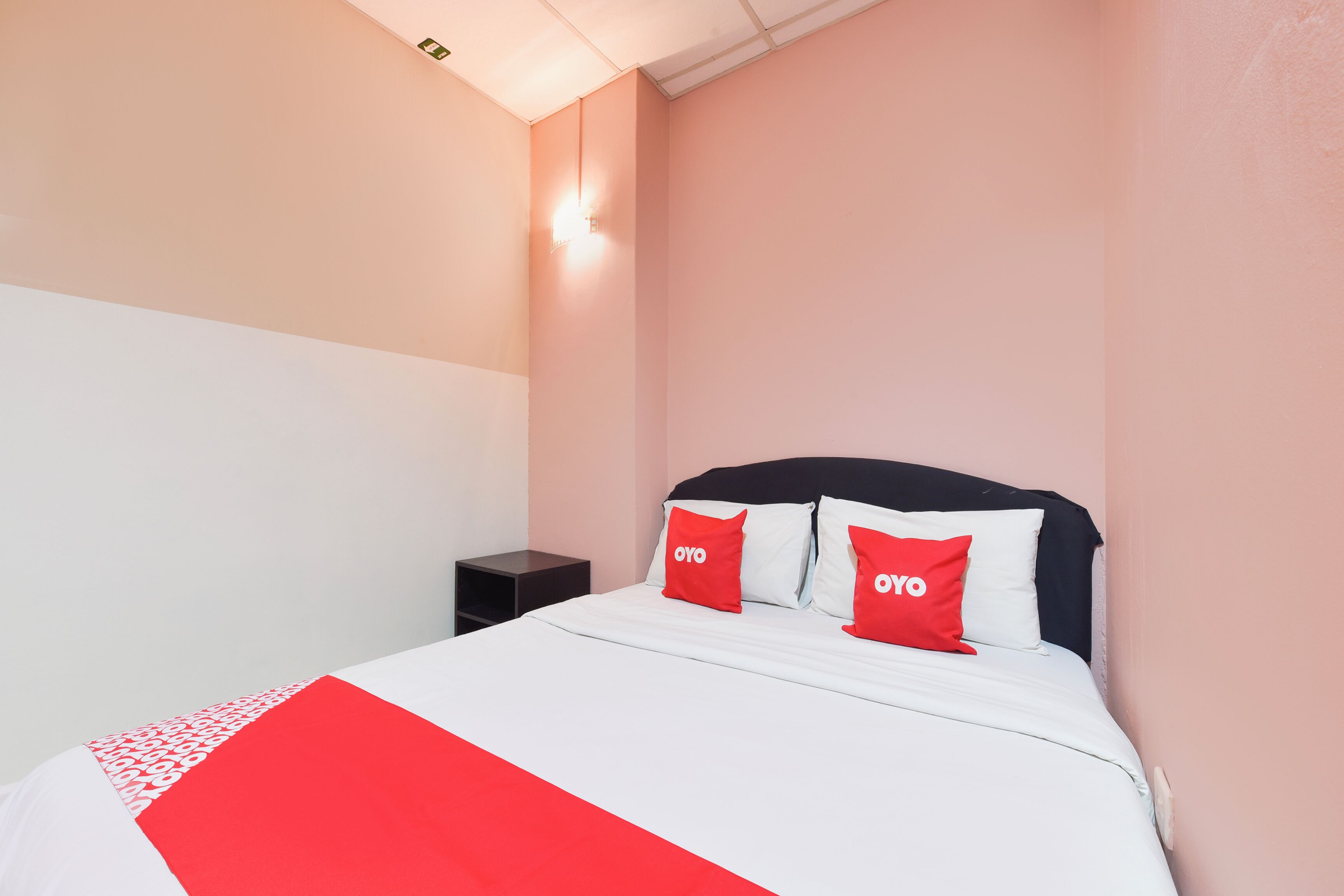Habitación de hotel con una cama, dos almohadas rojas con la marca "OYO", una mesita de noche y una lámpara fijada en la pared.