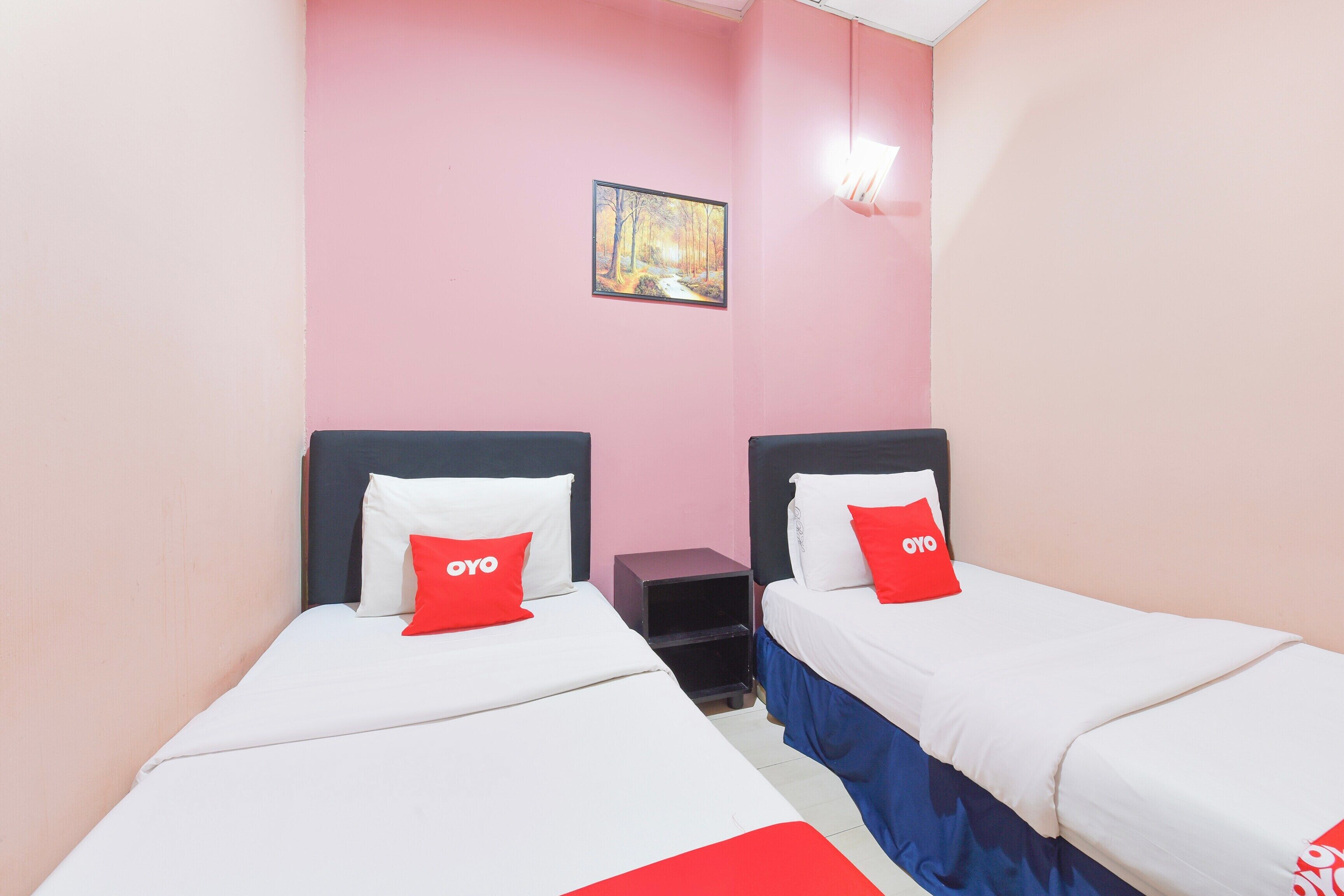 A room with two beds, each with a white mattress and a red pillow. The walls are pink, and there is a small painting on the wall.