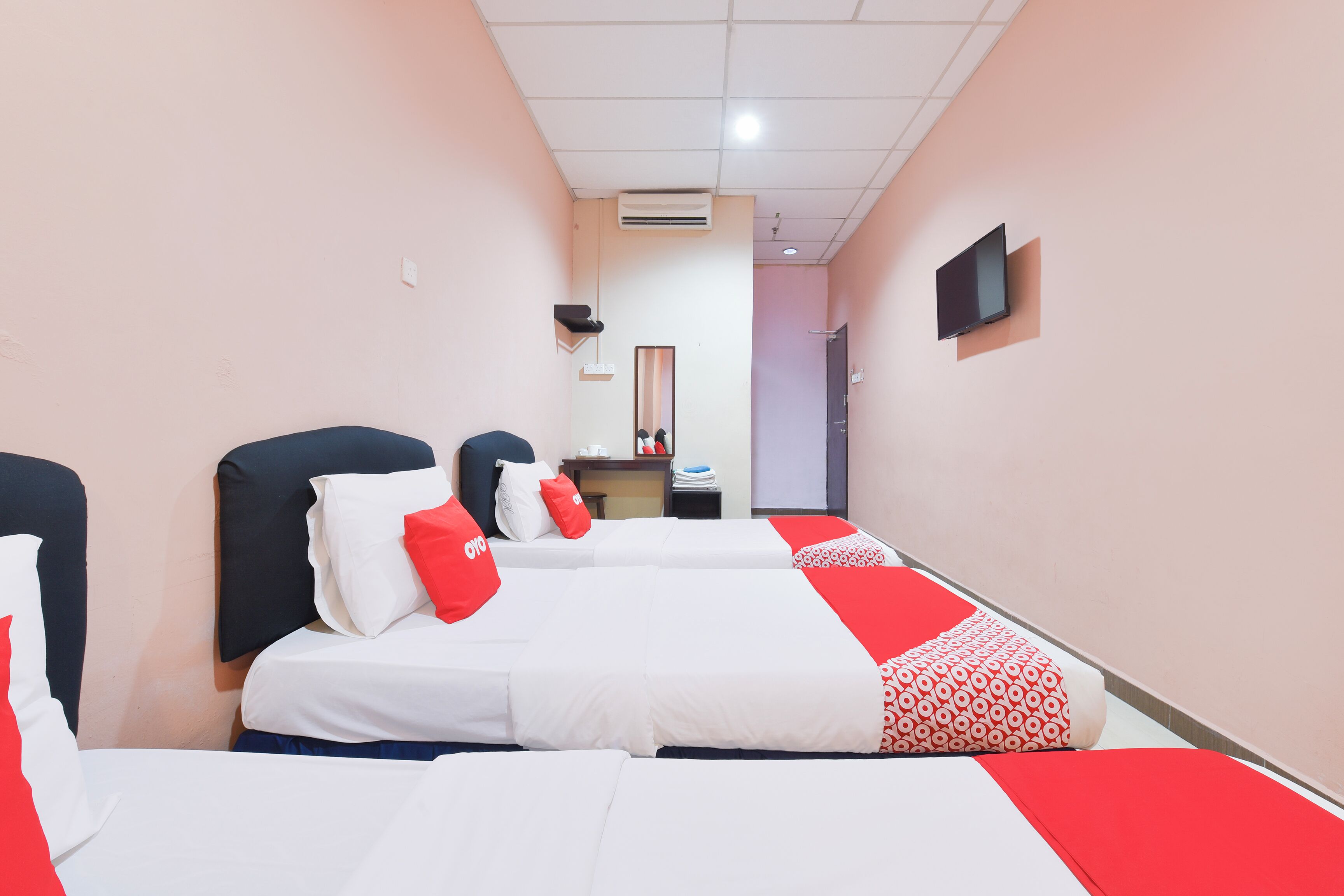 A hotel room with three beds, each with white bedding and red accents. There is a flat-screen TV mounted on the wall, an air conditioning unit on the ceiling, and a mirror above the desk.