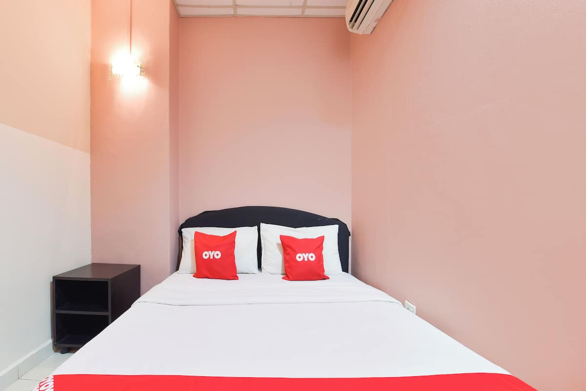 A neatly made bed with white bedding and two red pillows with "OYO" text, a small bedside table, and a wall-mounted air conditioner.
