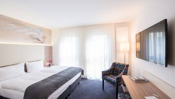 Quarto de hotel com cama, uma cadeira, escrivaninha e televisão.