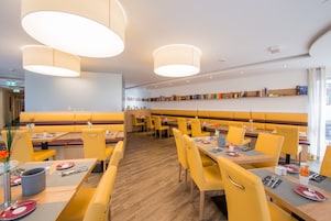 Un restaurante moderno con asientos y mesas amarillos, iluminación colgante y una estantería en el fondo.