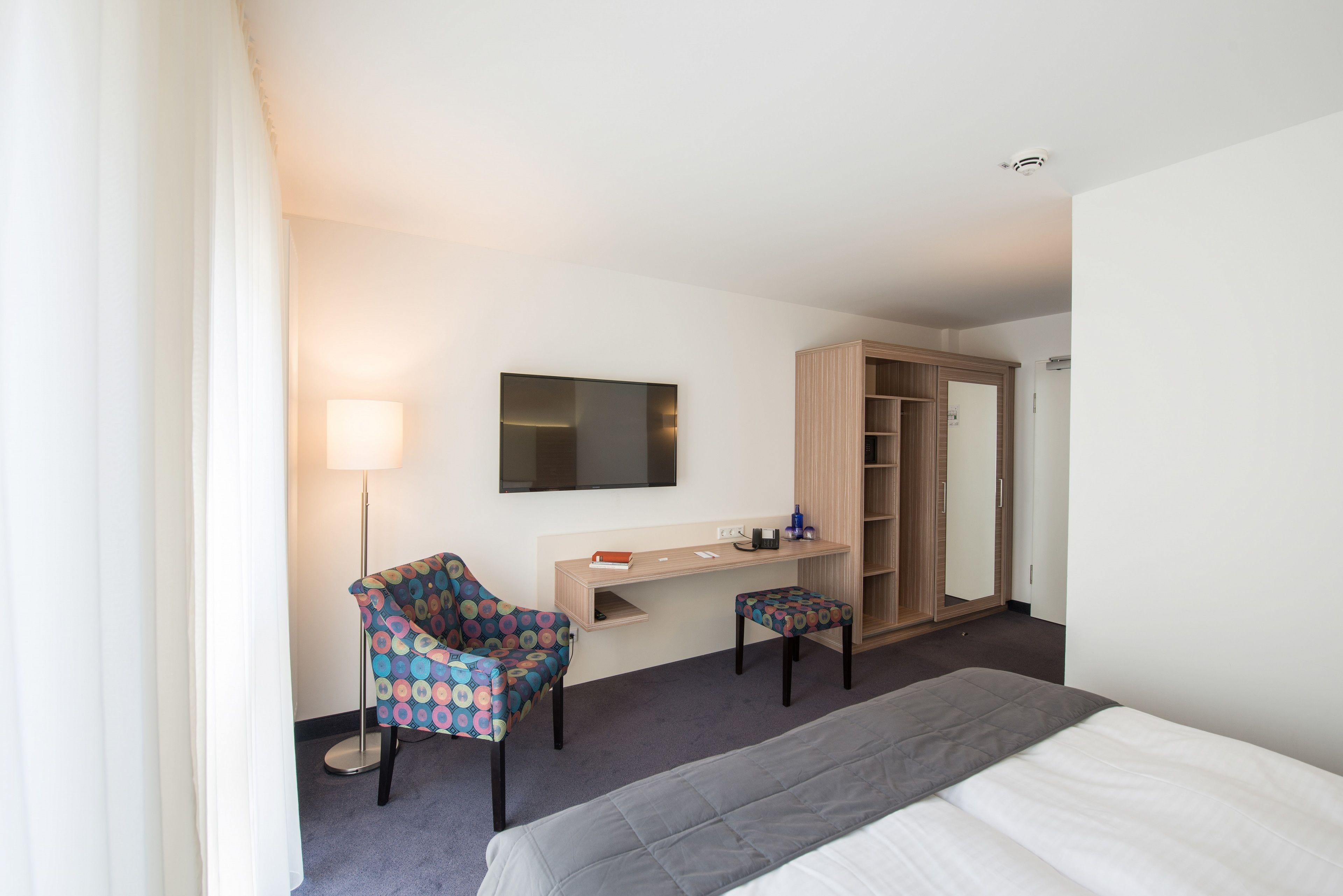 Ein Hotelzimmer mit Bett, Schreibtisch mit Stuhl, Kleiderschrank und einem an der Wand befestigten Fernseher.