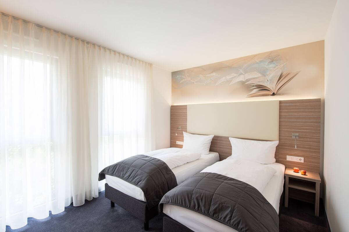 Una habitación de hotel con dos camas, un ventanal con cortinas translúcidas, una obra de arte colgada en la pared y una mesita de noche con lámpara.