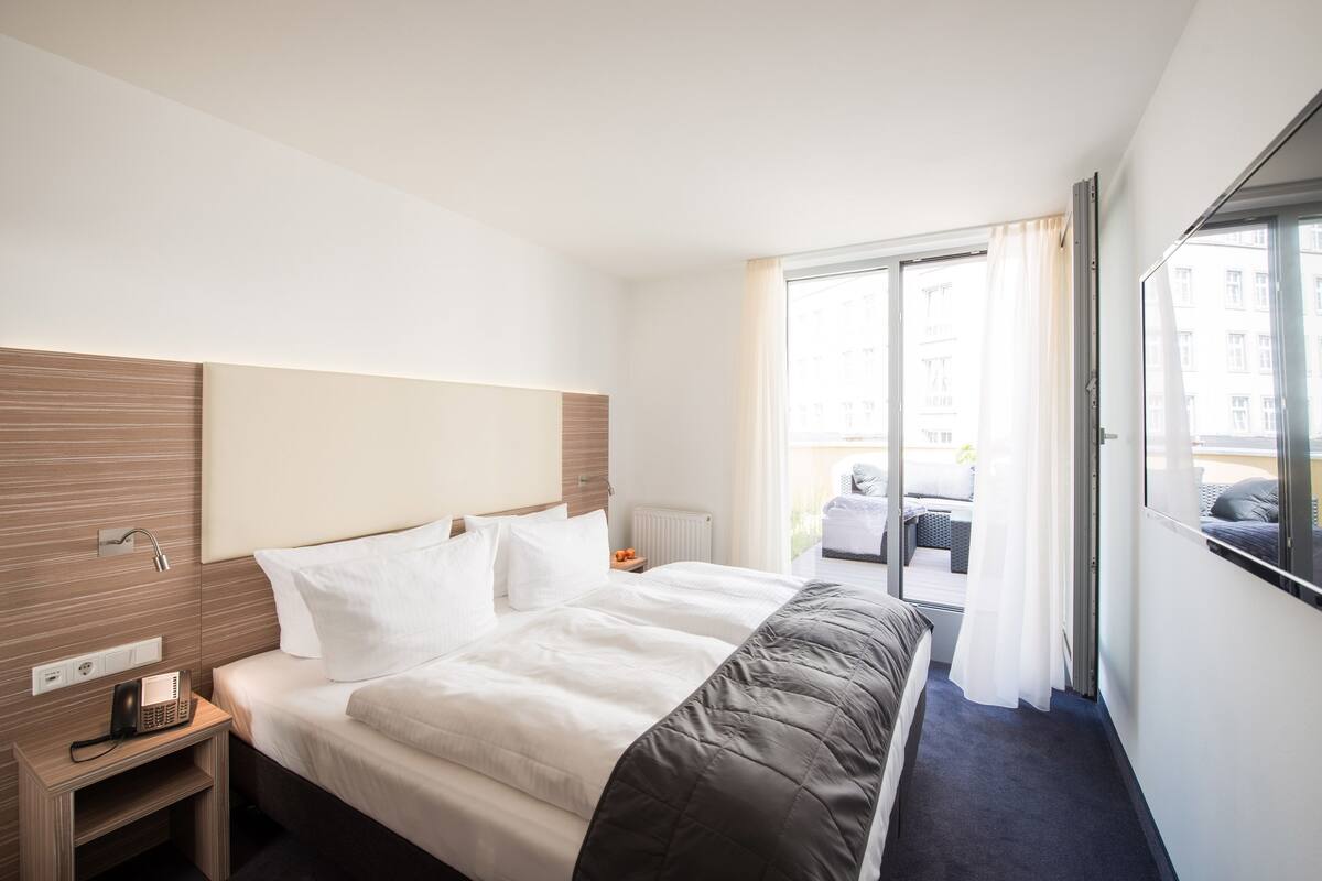 Habitación de hotel con una cama grande, teléfono en la mesita de noche, ventana con cortinas y vistas al balcón exterior.
