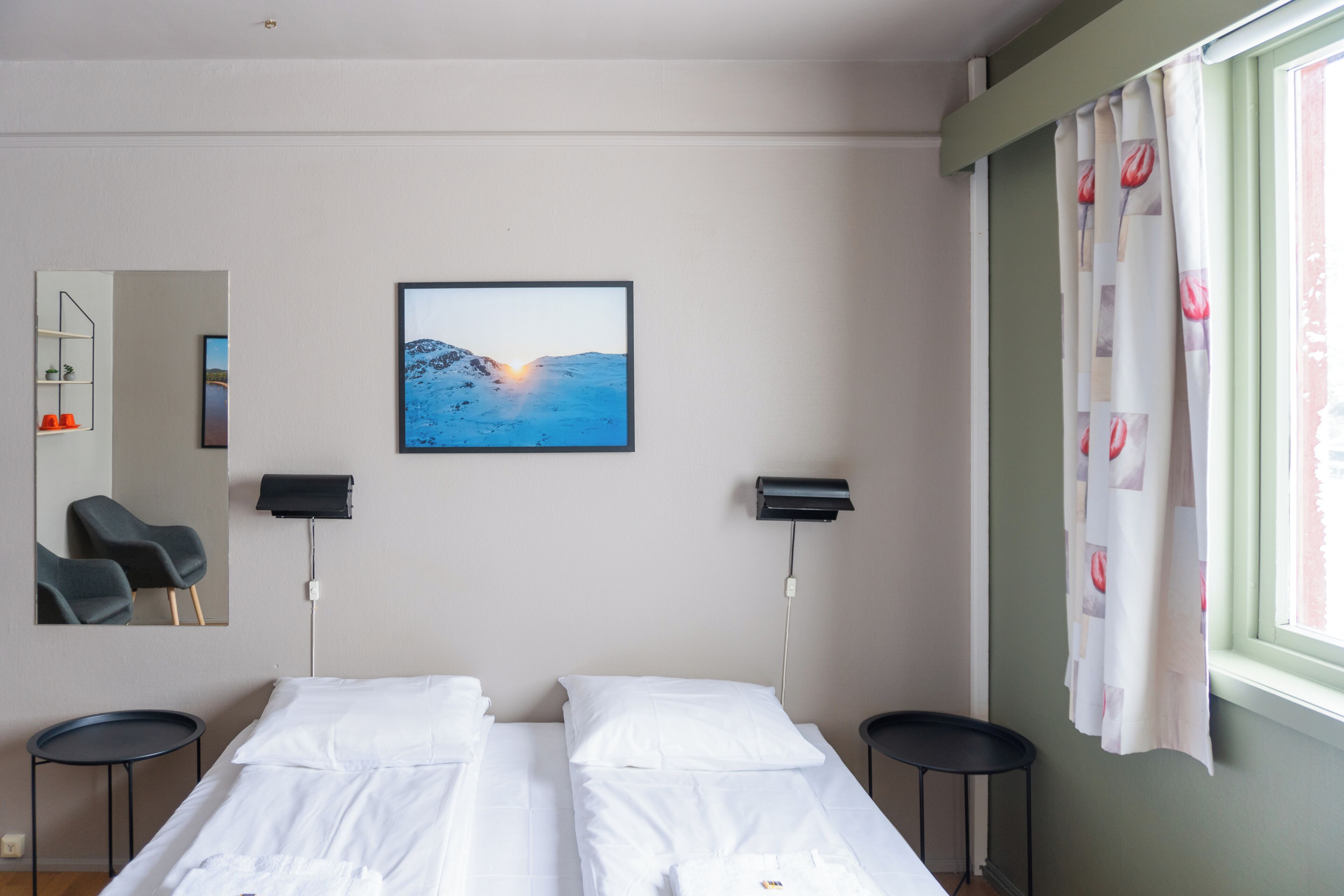Une chambre d’hôtel avec un lit, une chaise, un bureau, un miroir, un tableau au mur et une fenêtre avec des rideaux.