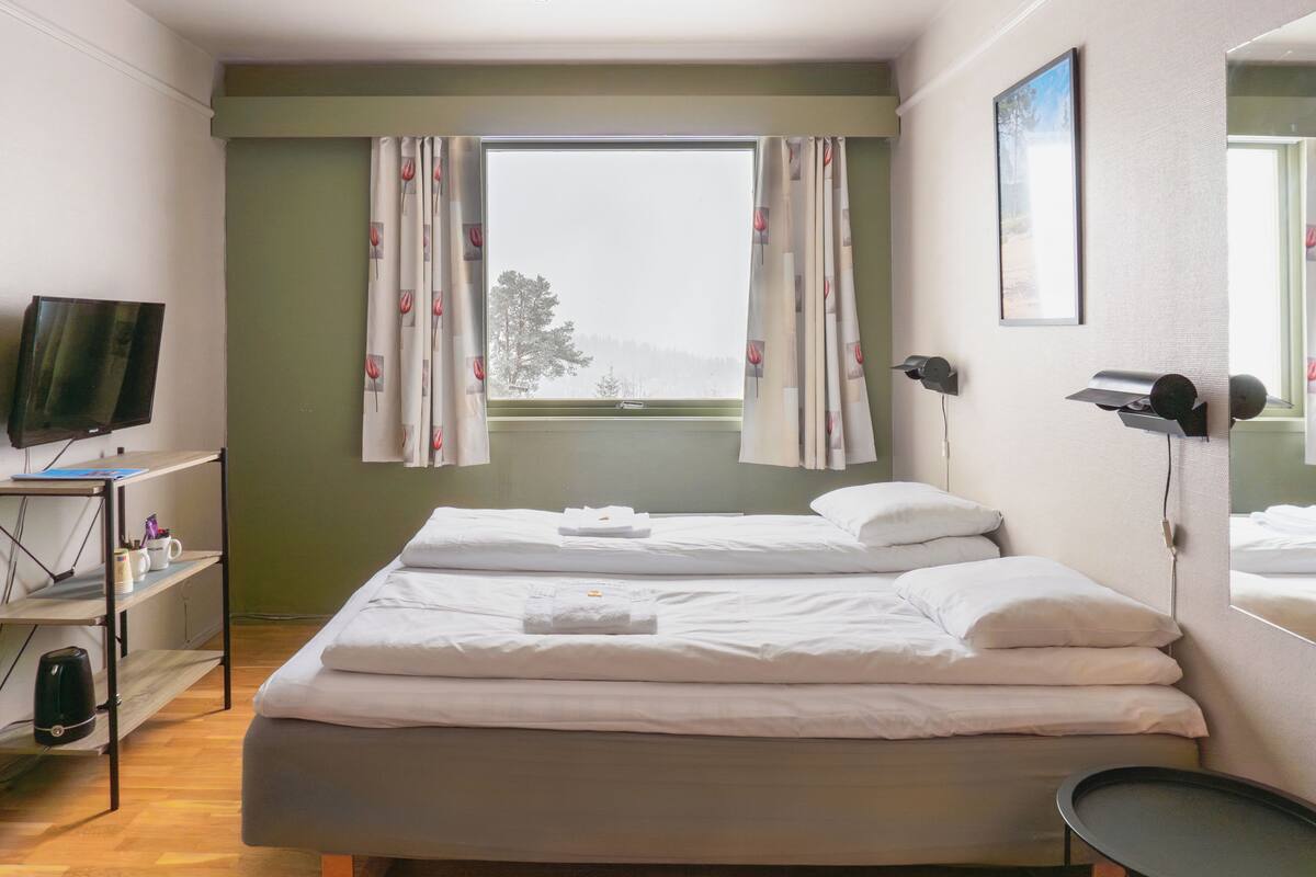 Habitación de hotel con cama, televisor, escritorio con ordenador y ventana con cortinas.