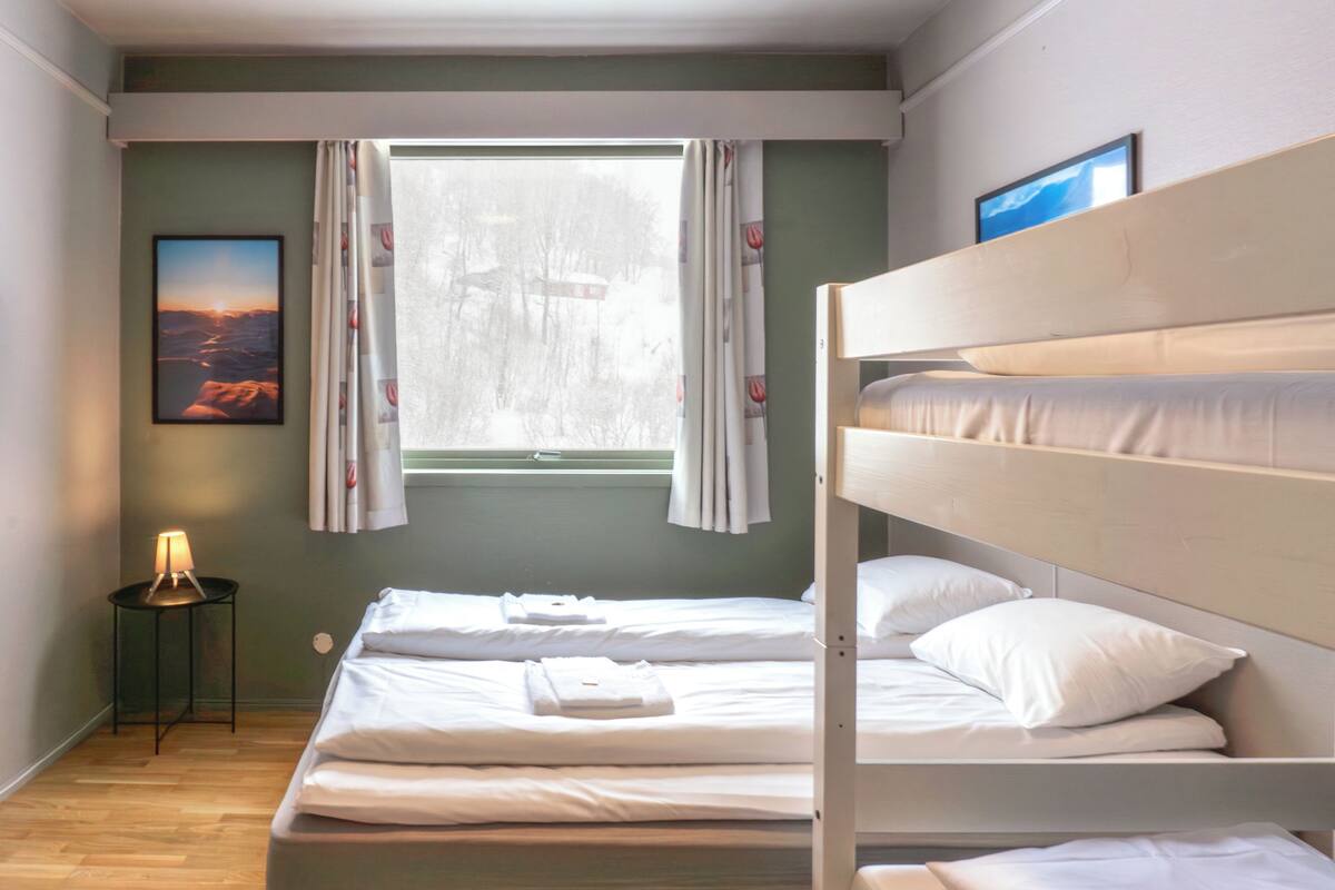 Habitación con literas, una ventana, un cuadro en la pared y una mesita con lámpara.