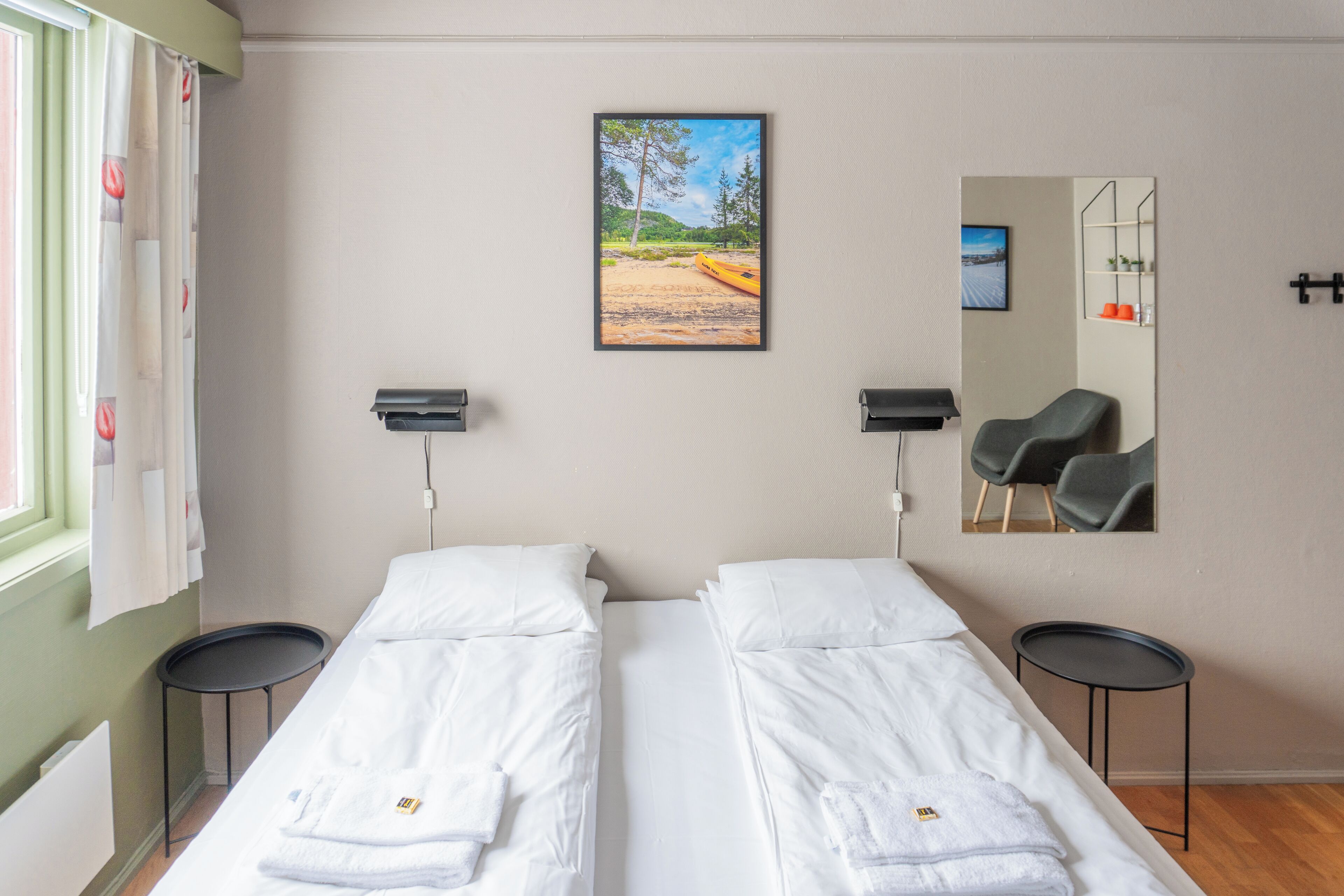 A hotel room with two single beds, a large window with curtains, a painting of a beach scene, a mirror, and two chairs.