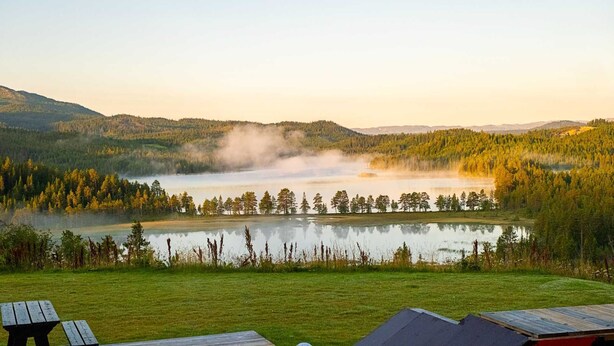 Une vue sur le lac, avec une terrasse en bois et des tables de pique-nique, le tout entouré d’une forêt dense et de collines.