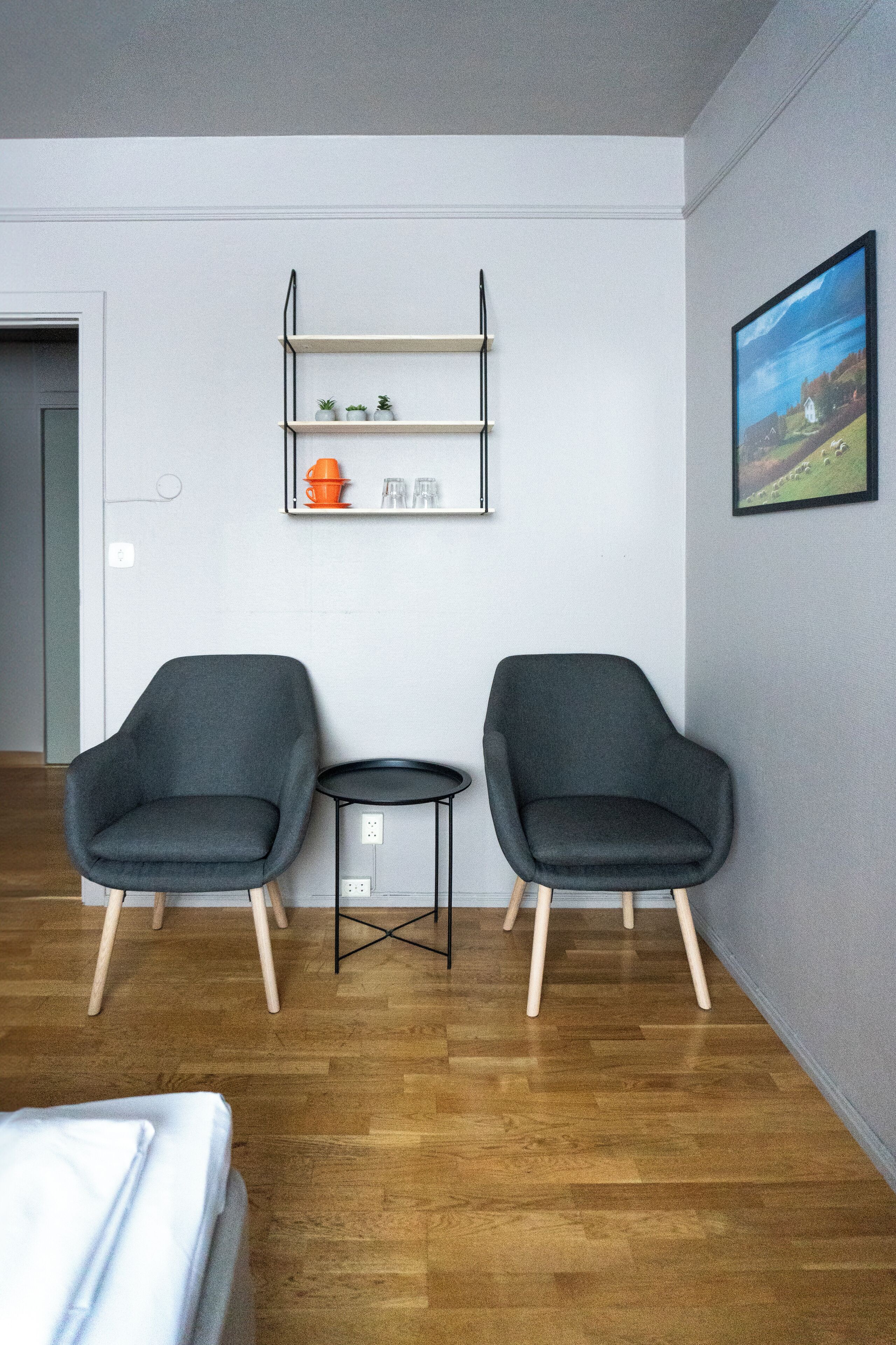 A room with two grey armchairs, a small table, and a shelf with decorative items.