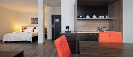 A modern hotel room with a bed, a desk, a chair, and a kitchenette.