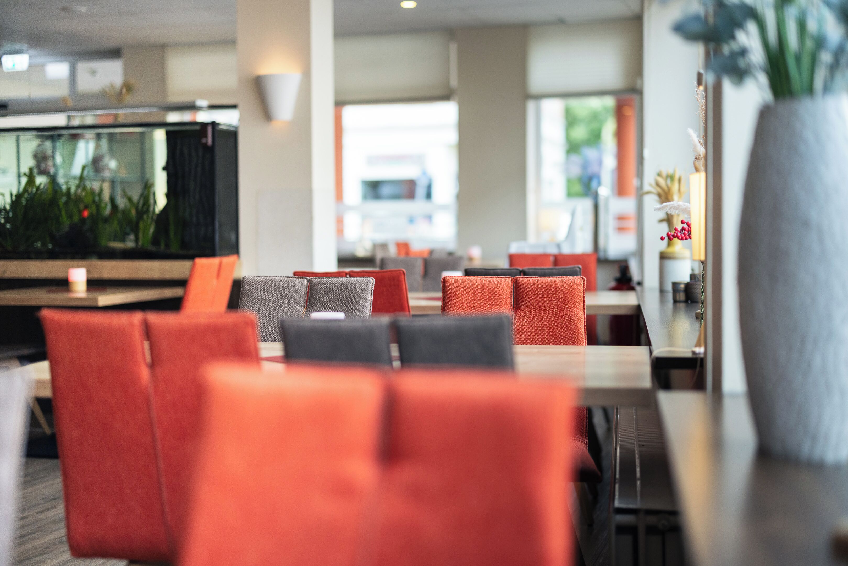 Un restaurant moderne avec des chaises rembourrées de couleur rouge et grise, un grand aquarium et un grand vase contenant des plantes séchées.