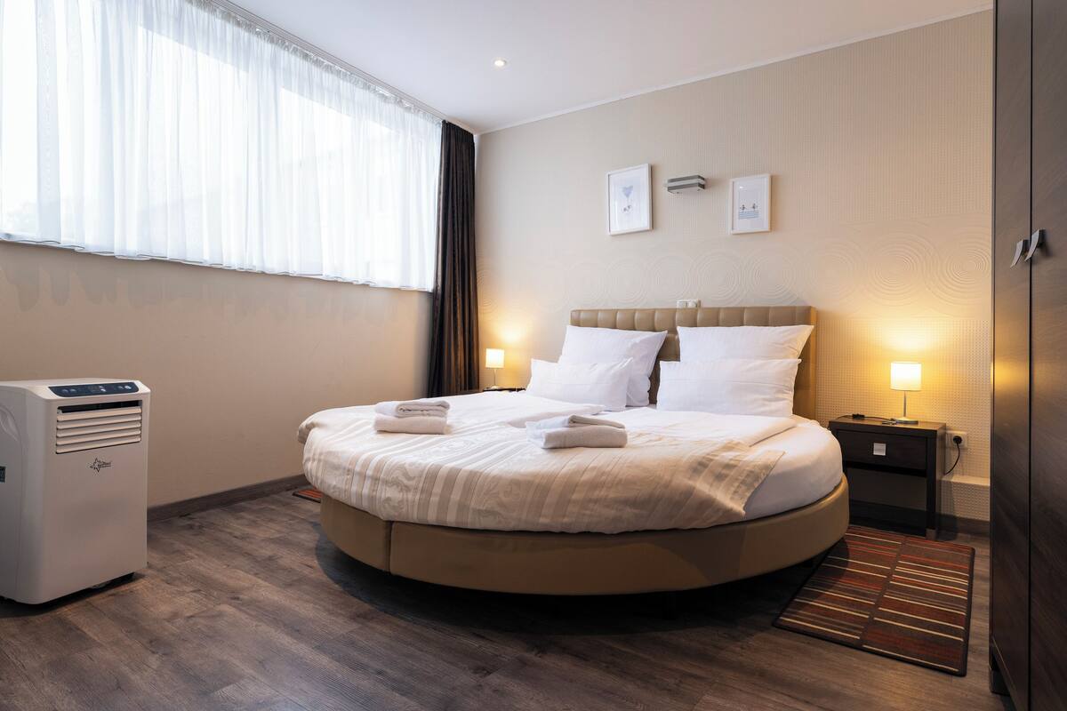 A hotel room with a round bed, a bedside table with a lamp, a heater, and a window with sheer curtains.