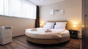 Habitación de hotel con cama redonda, mesita de noche con lámpara, calefactor y ventana con cortinas de gasa.