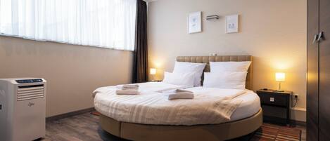 A hotel room with a round bed, a bedside table with a lamp, a heater, and a window with sheer curtains.