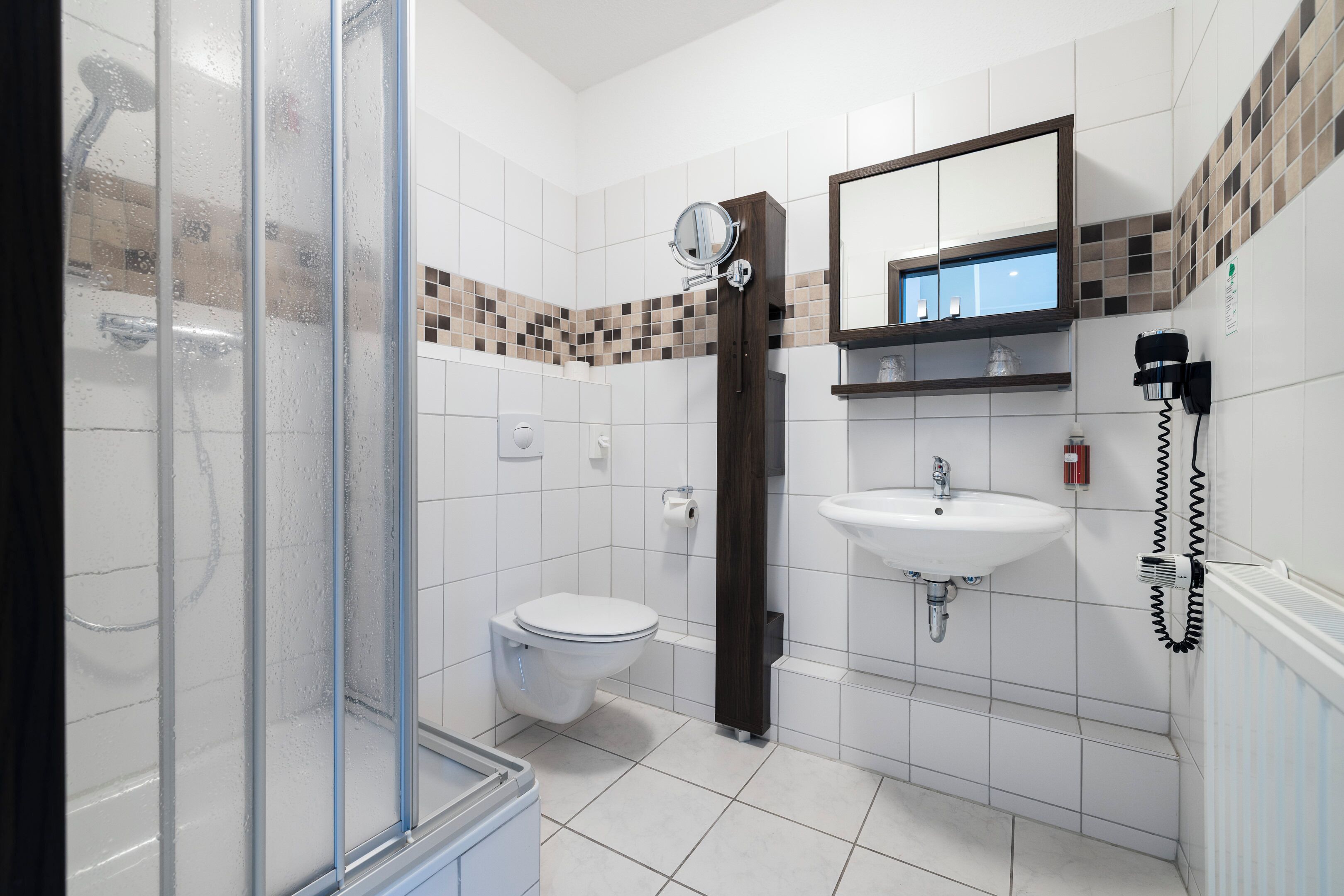 Une salle de bain moderne avec une douche à l’italienne en verre, des toilettes, un lavabo avec un miroir au-dessus et un sèche-cheveux fixé au mur.