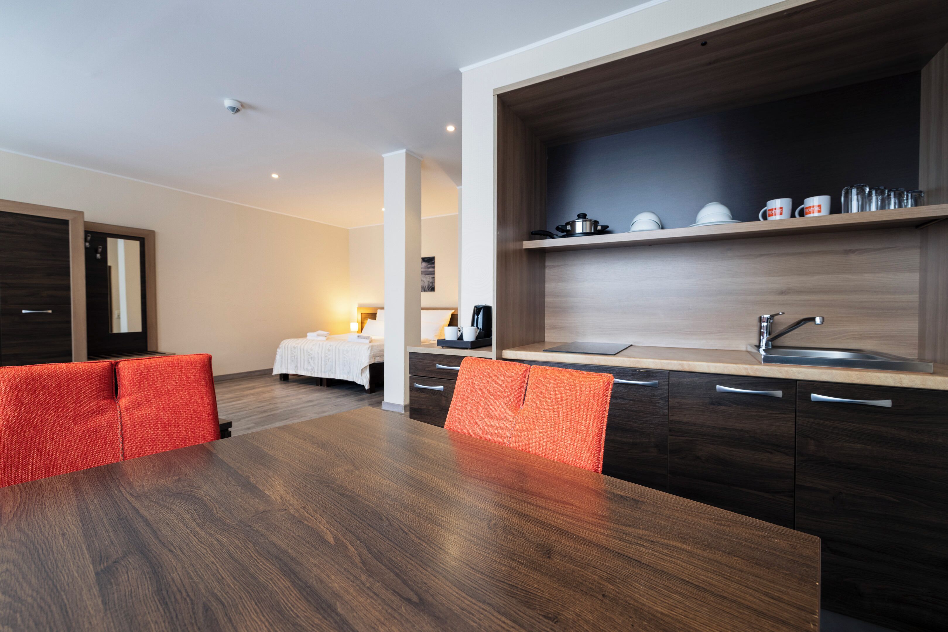 A modern hotel room with a wooden dining table, red upholstered chairs, a kitchen area with a sink, and a bed with a white duvet.
