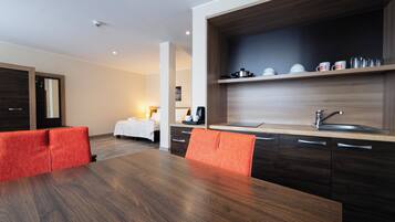 A modern hotel room with a wooden dining table, red upholstered chairs, a kitchen area with a sink, and a bed with a white duvet.