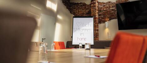 A conference room with a whiteboard displaying 'Lich Willkommen im home' message, a wooden table, and a brick wall.