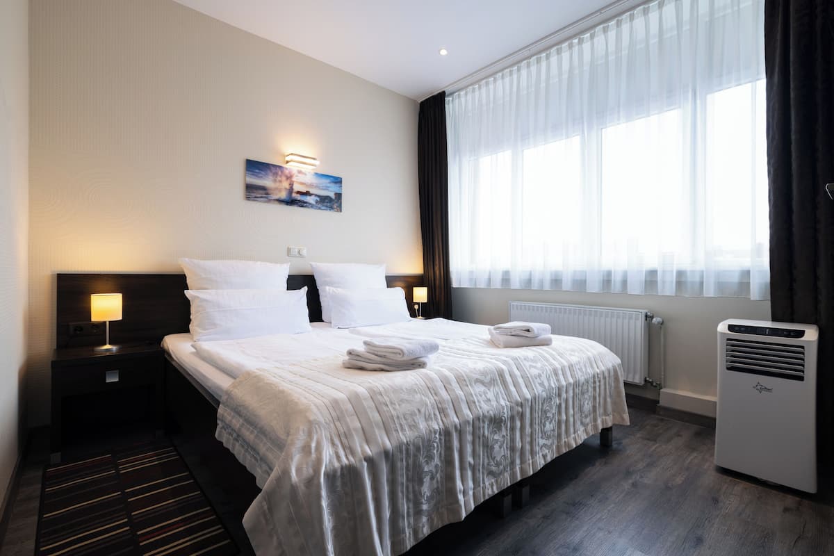 A hotel room with a large bed, a nightstand with a lamp, a radiator, and a window with curtains.
