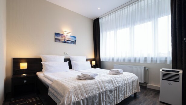 Habitación de hotel con una cama grande, una mesita de noche con lámpara, un radiador y una ventana con cortinas.
