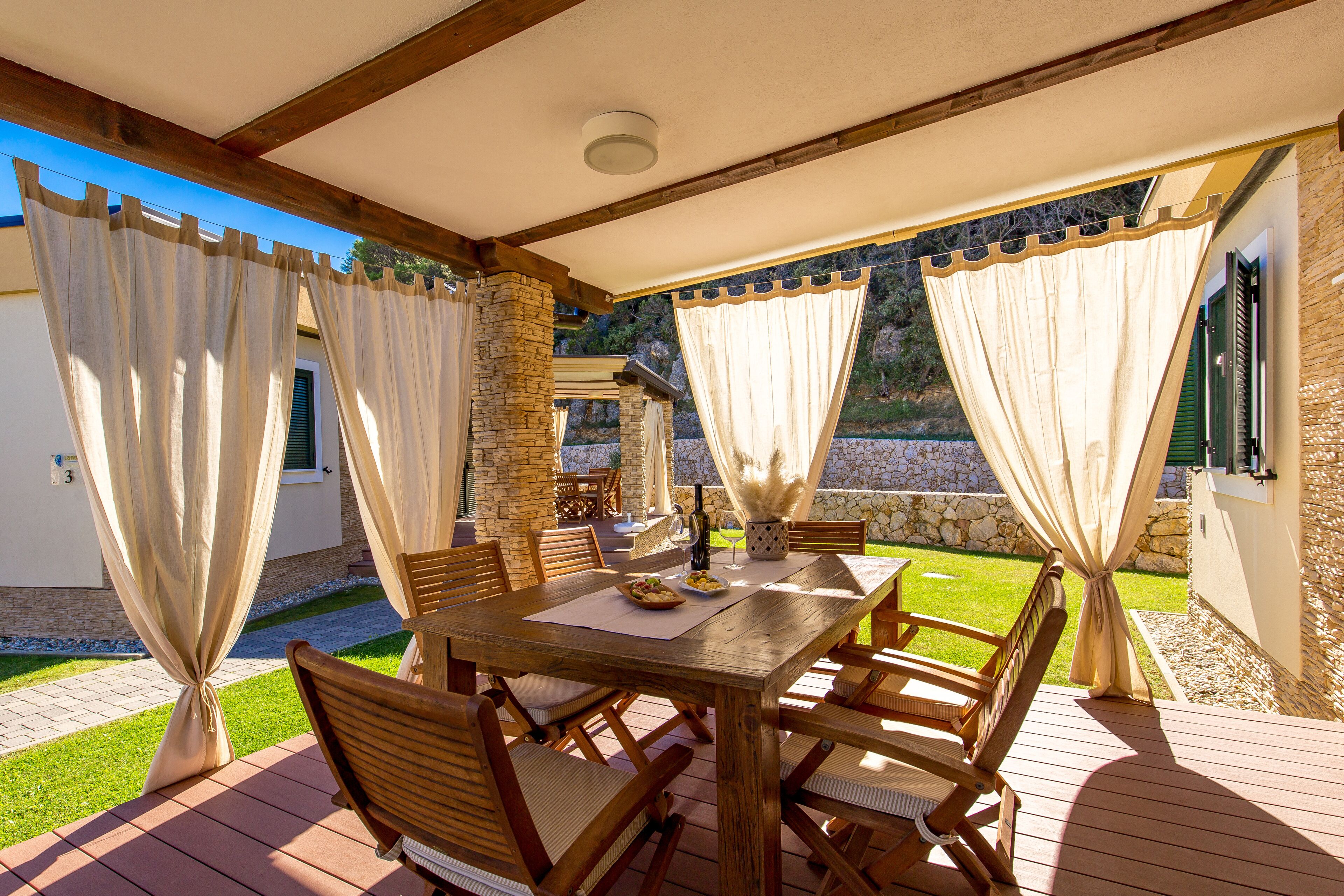 An outdoor patio with a wooden table and chairs, stone pillars, and a view of a stone wall and greenery.