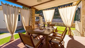 An outdoor patio with a wooden table and chairs, stone pillars, and a view of a stone wall and greenery.