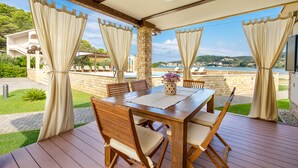 An outdoor patio with a wooden table and chairs, beige curtains, and a view of the sea.