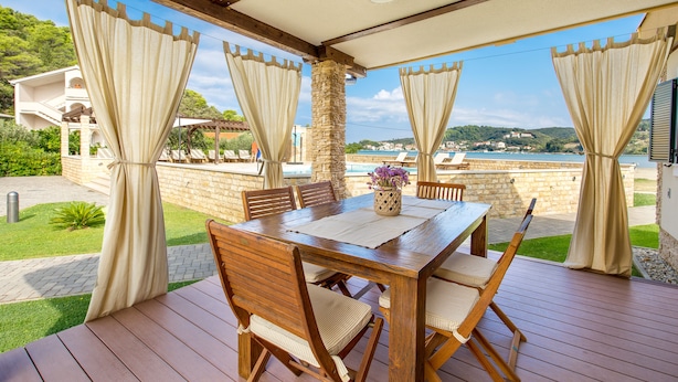 An outdoor patio with a wooden table and chairs, beige curtains, and a view of the sea. - Lando Resort (Rab)
