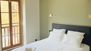 Una cama bien hecha con sábanas y toallas blancas, un cabecero oscuro y vistas a un edificio de piedra a través de una puerta corrediza.