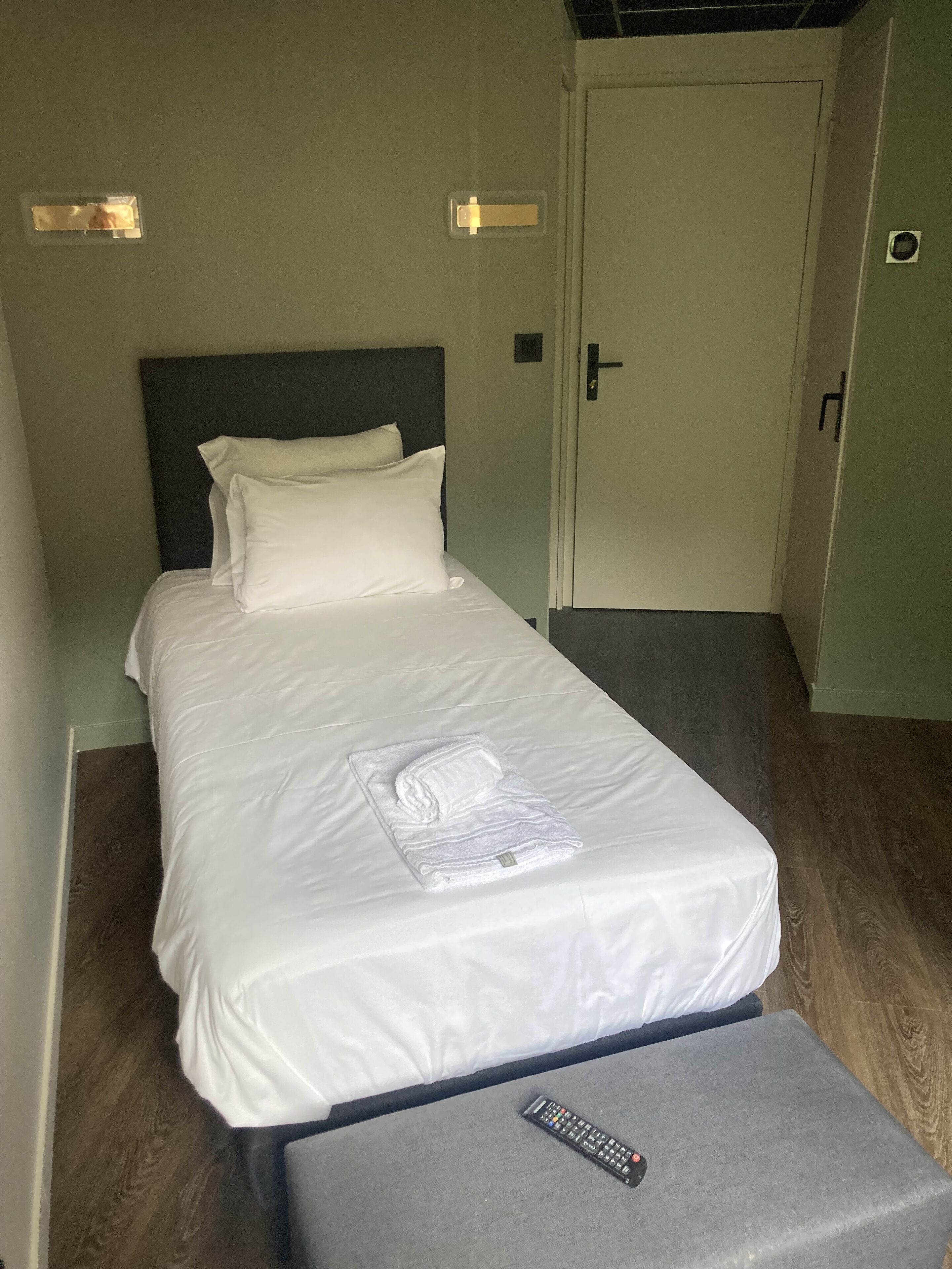 A single bed with white linens, a headboard, and a remote control on a small table.