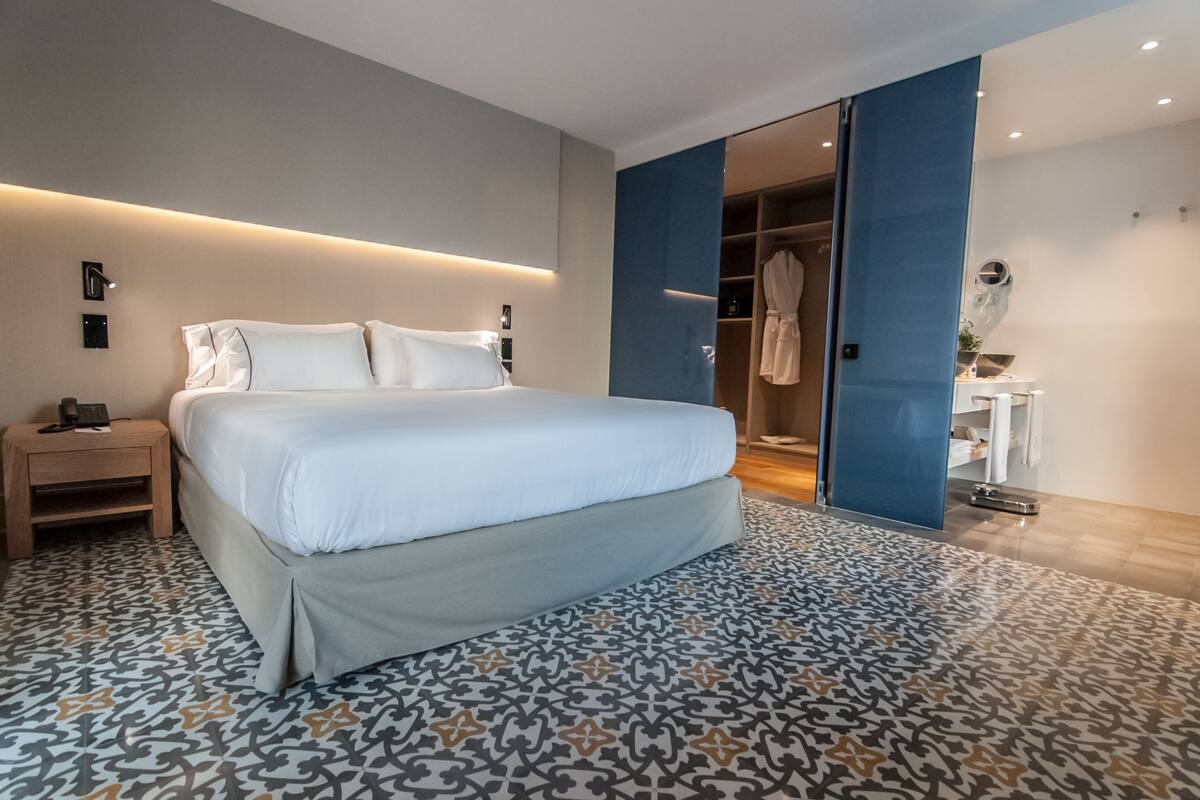 A modern hotel room with a large bed, a patterned rug, a walk-in closet, and a bathroom with a mirror and sink.