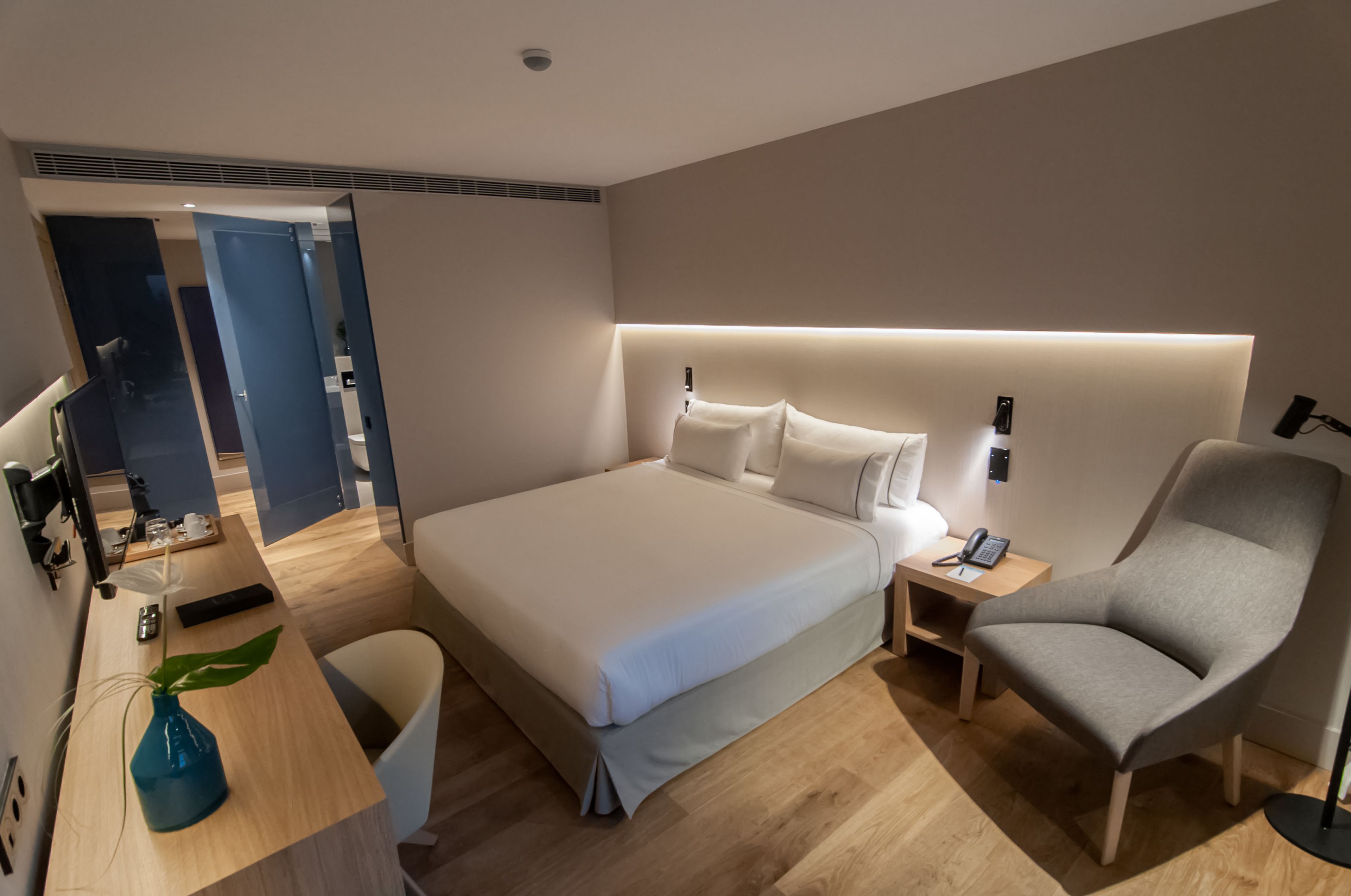 Habitación de hotel moderna con una cama grande, mesitas de noche, una silla y un escritorio con una planta.