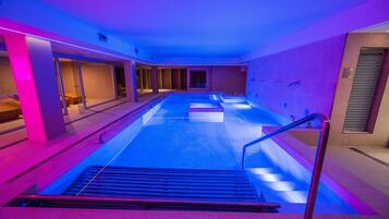 A modern indoor swimming pool with a glass wall on one side, a metal railing, and a tiled floor.