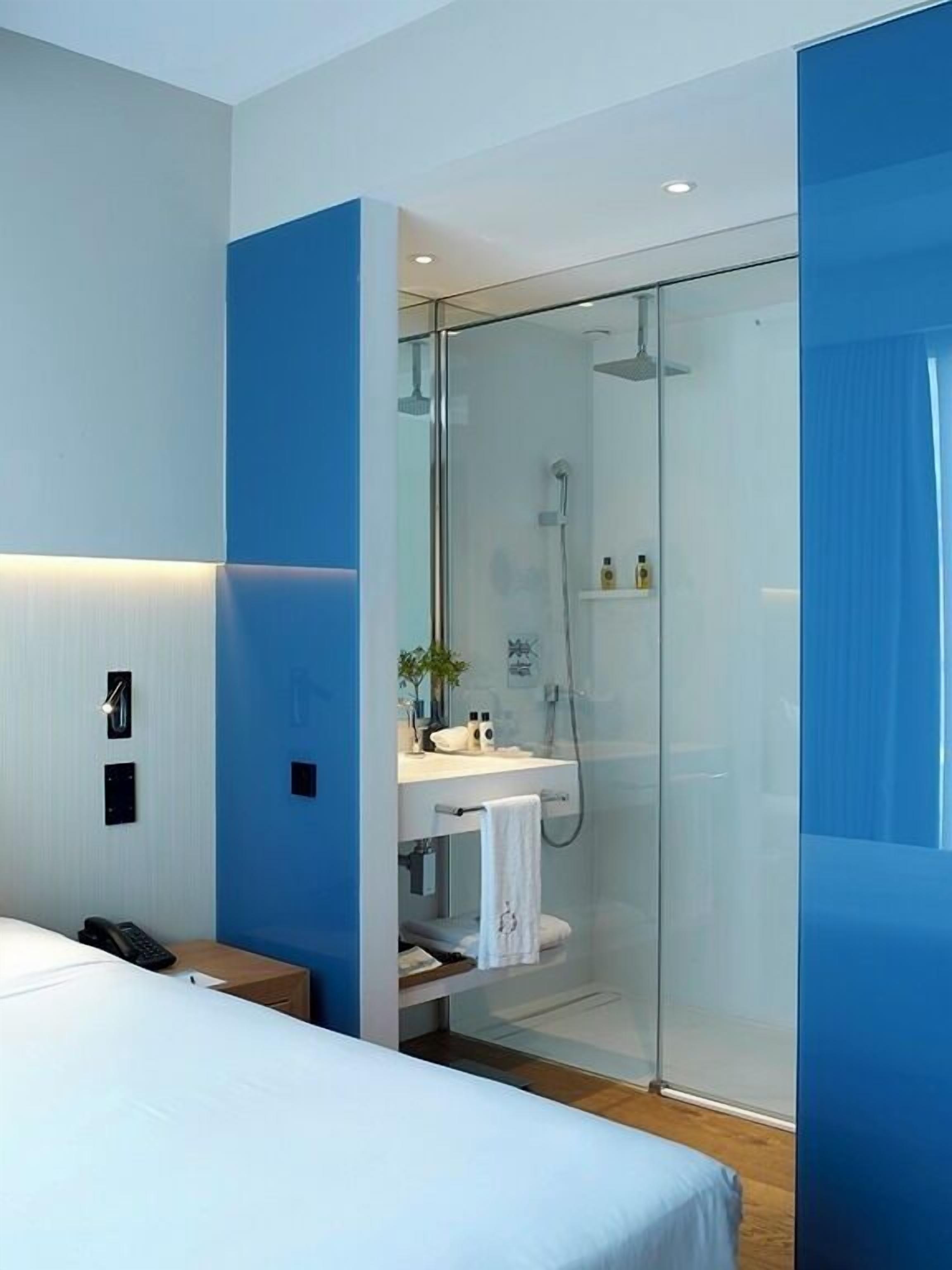 Una habitaci ón de hotel moderna con ducha de vidrio, una mesita de noche, una cama y un escritorio pequeño con un teléfono.