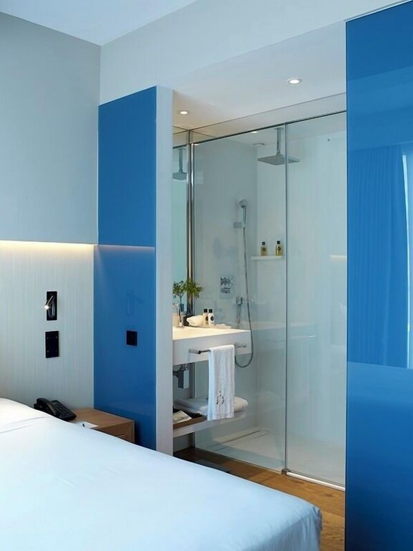 A modern hotel room with a glass-enclosed shower, a bedside table, a bed, and a small desk with a telephone.